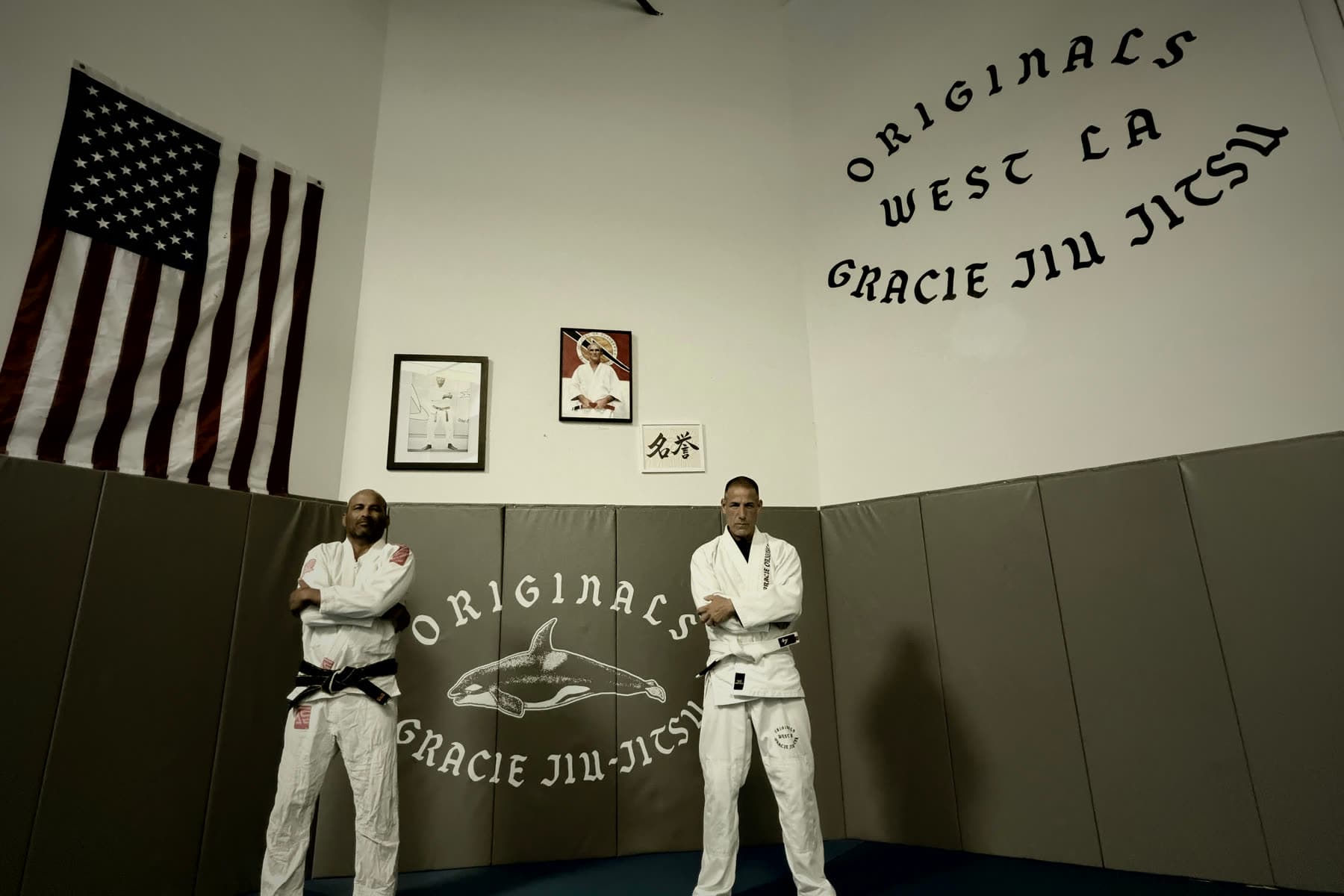 Two practitioners in gis standing on the mats beneath the lineage wall at Originals Gracie Jiu-Jitsu